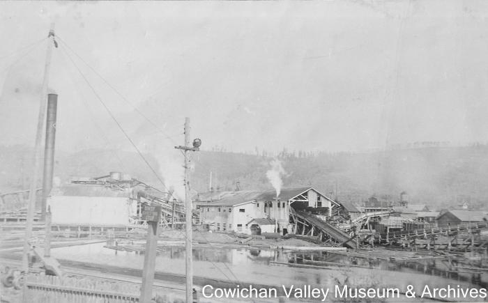 Looking north towards Hill 60, and showing the mill with logs on the log deck, the boiler shack and the mill pond.