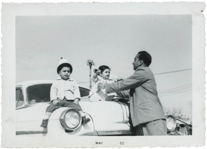 [Photo of two young children sitting on a car with an unidentified man holding them]